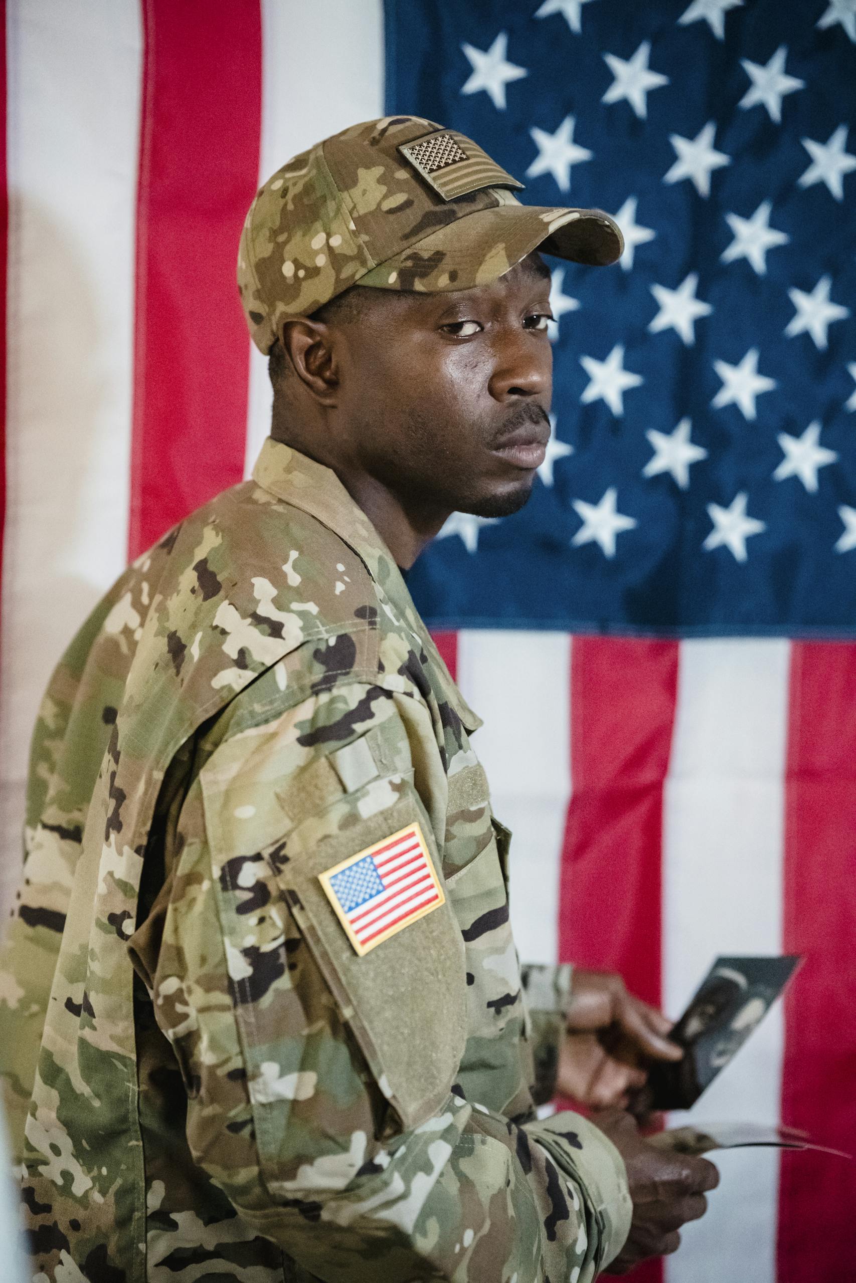 A soldier in military uniform stands with an American flag backdrop, holding a photo.